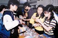 校 慶 義 賣 攤 位 上 ， 德 文 系 同 仁 仔 細 地 向 詢 問 的 顧 客 解 說 展 示 物 品 的 使 用 方 法 。